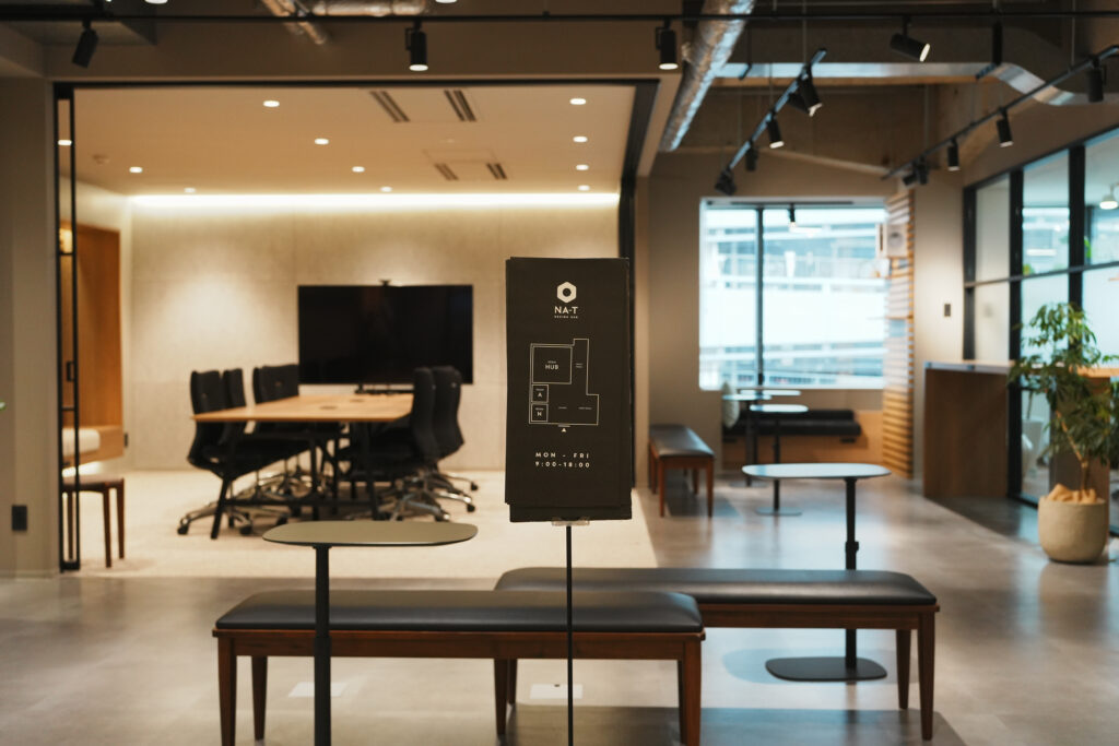 NA-T DESIGN HUB(ナッティデザインハブ)内のショールーム及び会議室「HUB」の写真。手前に施設内の案内図があり、奥には大型モニターを備えた間仕切りを変更可能な会議室が見える。